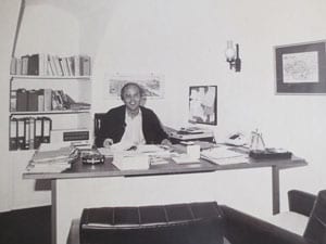 Rechtsanwalt-Klaus-Hafen-im-Jahr-1977 Rechtsanwalt sitzt am Schreibtisch im Vordergrund sind zwei Stühle, an der Wand hängt ein Bild, im Hintergrund steht ein Regal.